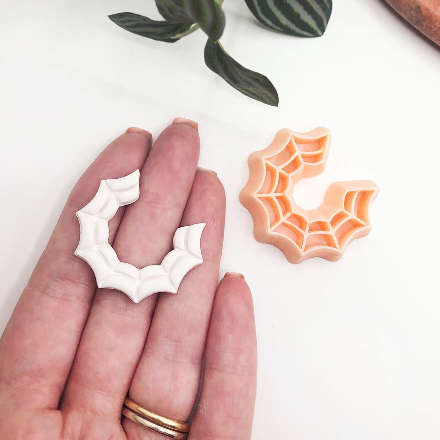 A hand holds a small white bat-shaped polymer clay cut out with a peach-colored spiderweb-patterned ring-shaped accessory lies beside it on a white surface with blurred green leaves in the background.