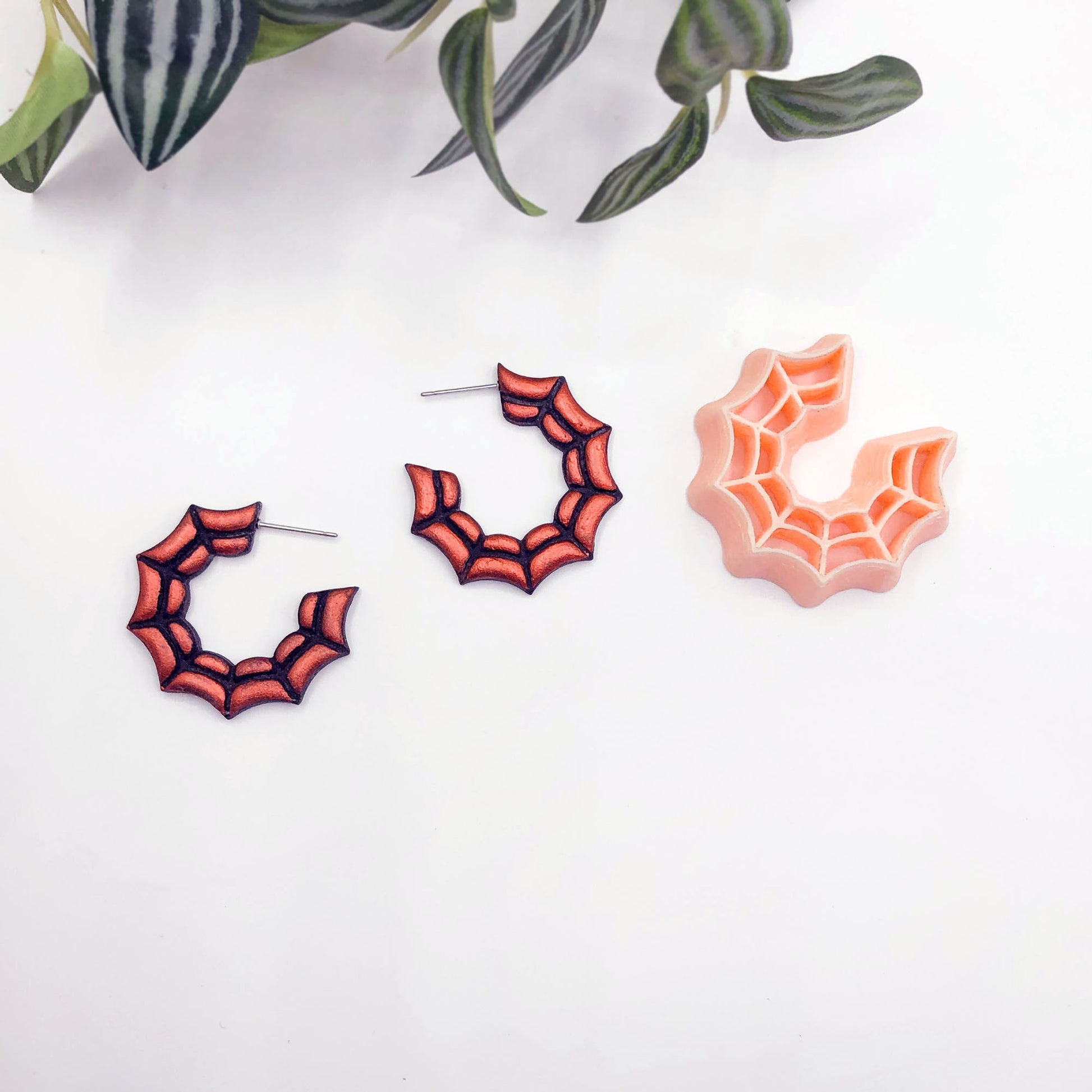 Two orange and black spiderweb hoop earrings displayed on a white surface with green leaves at the top, alongside a peach-colored spiderweb-shaped cookie clay cutter.