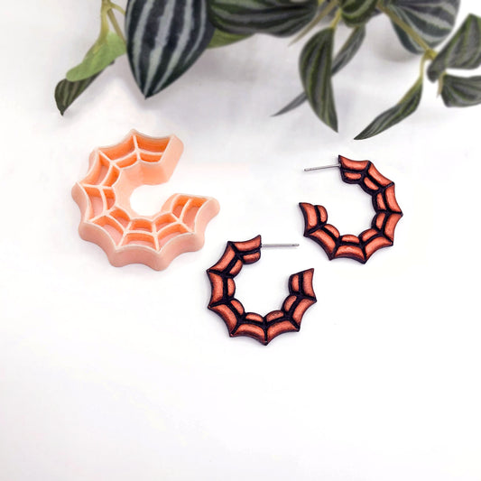 Spiderweb hoop earrings in orange and black, shown with a matching peach-colored spiderweb-shaped clay cutter on a white background with green plant leaves in the corner.
