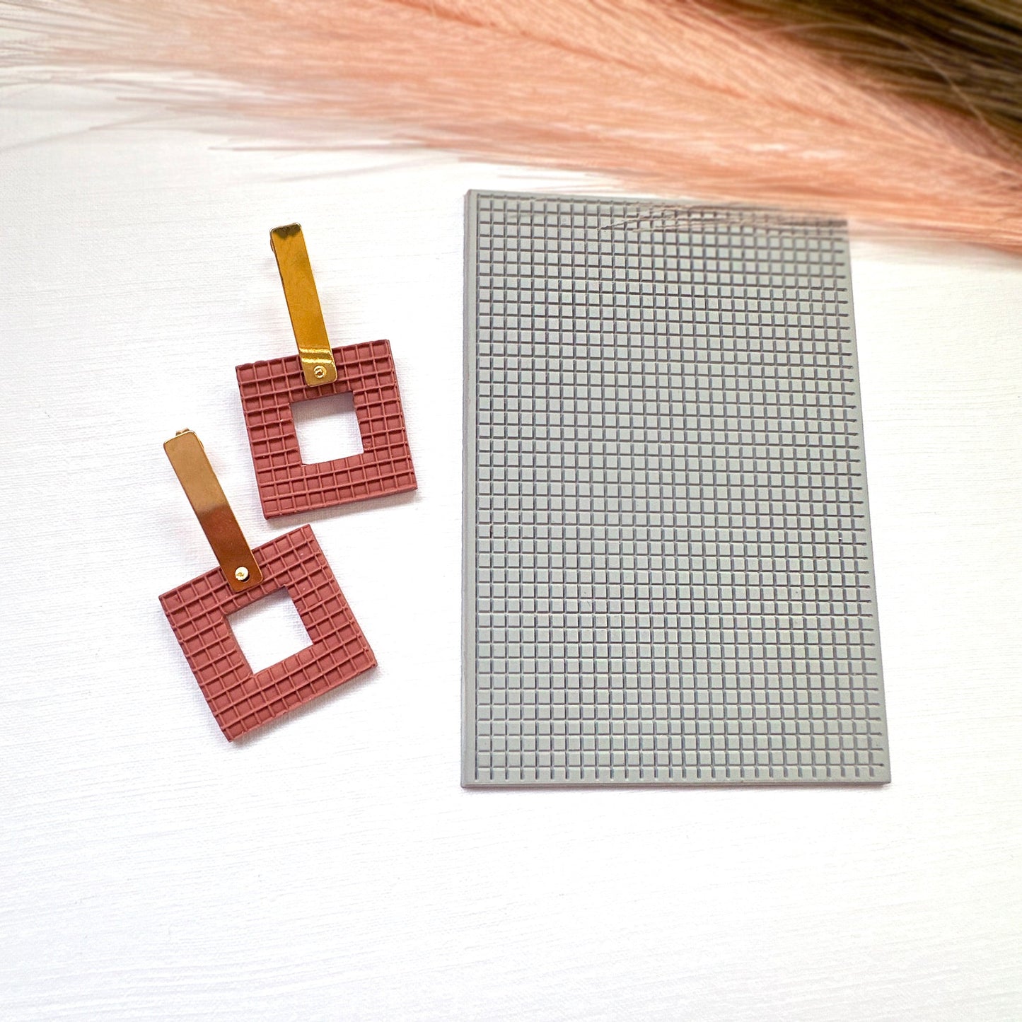 The picture shows a pair of brick coloured square earrings made from polymer clay, using the square plaid texture mat they are sat on. They are made with the square donut clay cutter.