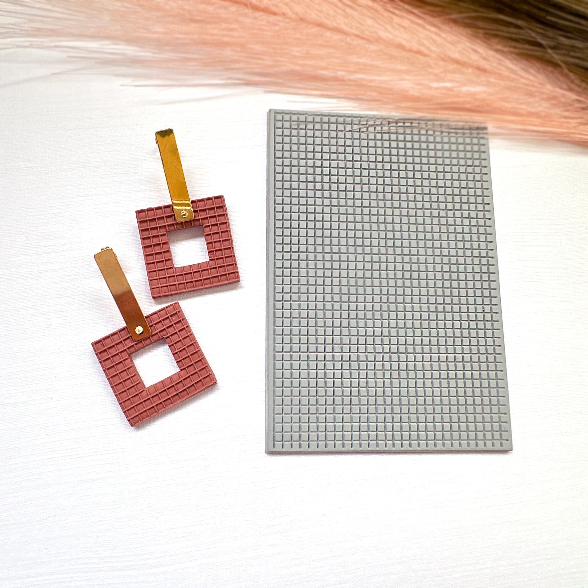 The picture shows a pair of brick coloured square earrings made from polymer clay, using the square plaid texture mat they are sat on. They are made with the square donut clay cutter.