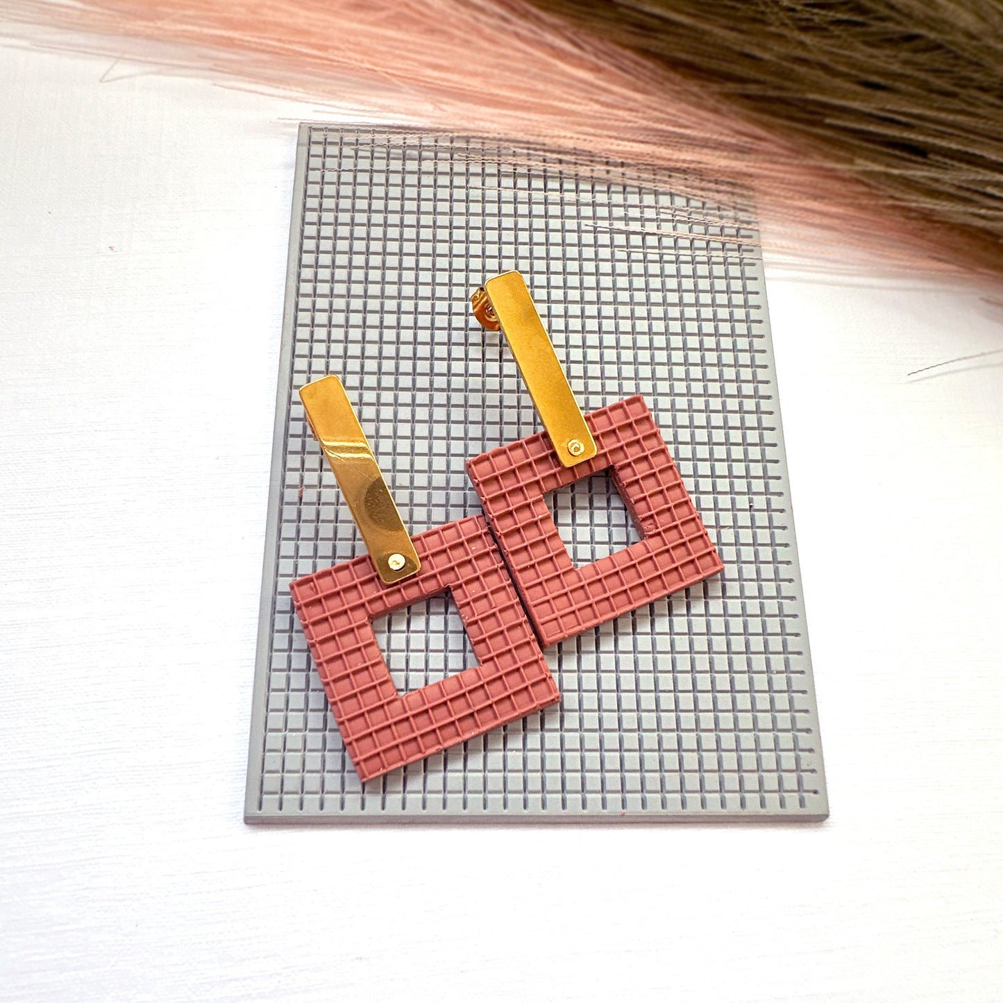The picture shows a pair of brick coloured square earrings made from polymer clay, using the square plaid texture mat they are sat on. They are made with the square donut clay cutter.