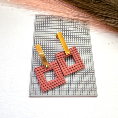 The picture shows a pair of brick coloured square earrings made from polymer clay, using the square plaid texture mat they are sat on. They are made with the square donut clay cutter.