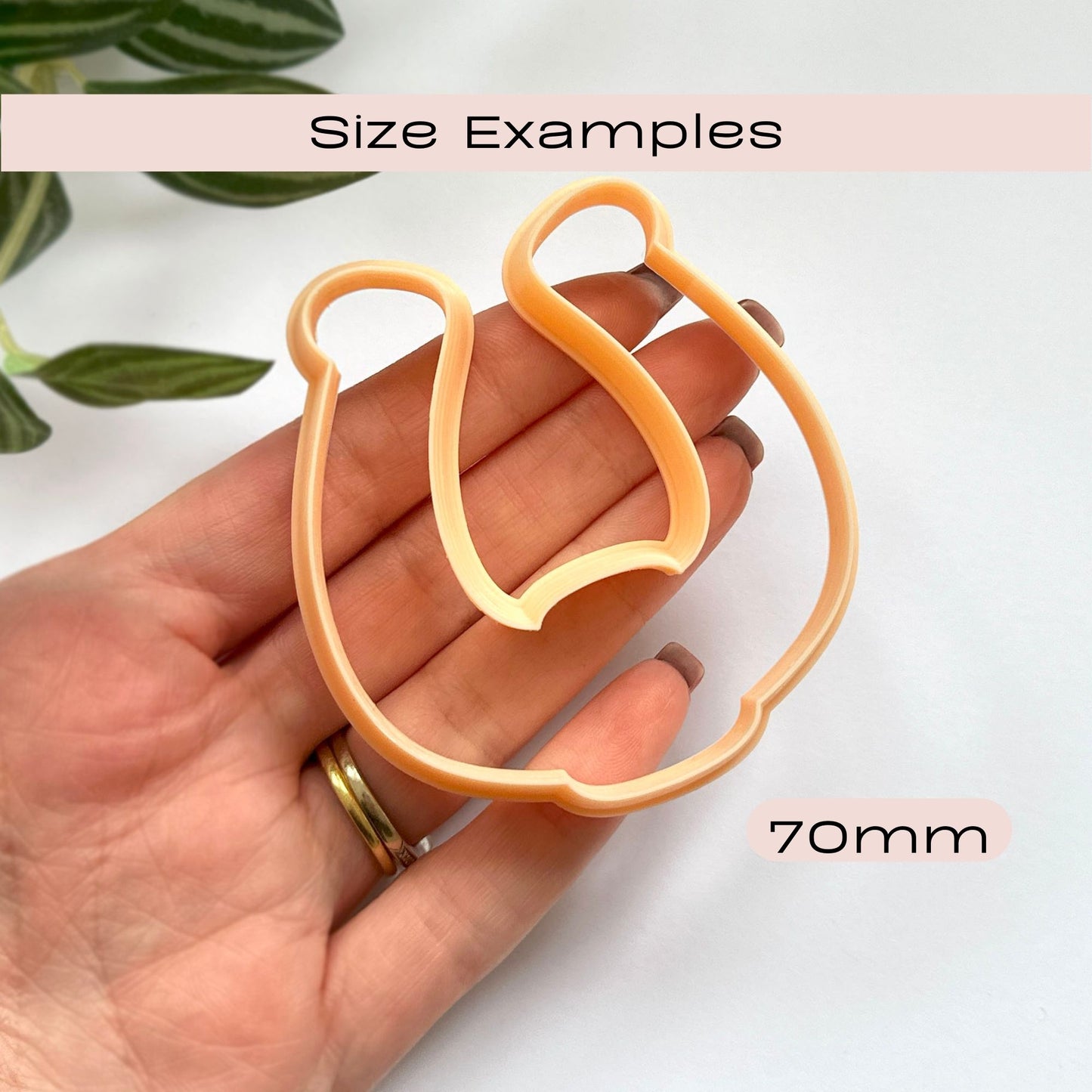 A hand holds a peach-colored cookie cutter shaped like a horseshoe, with text reading 'Size Examples' and '70mm' on a white background with a small plant leaf in the corner.