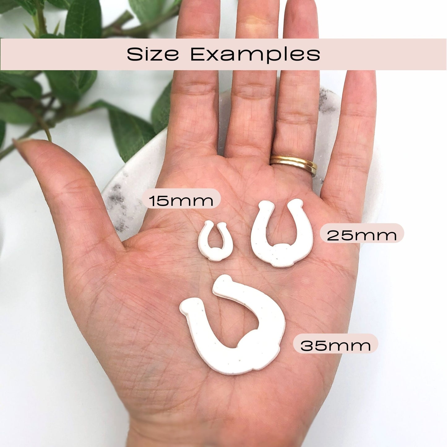 A hand holding three white horseshoe-shaped polymer clay cut outs labeled 15mm, 25mm, and 35mm, with a 'Size Examples' banner at the top to show scale.