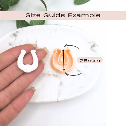 A size guide example showing a small horseshoe-shaped clay cut out in white held in a hand next to a matching horseshoe-shaped clay cutter in peach, with arrows and text indicating a 25mm size on a white marble surface.