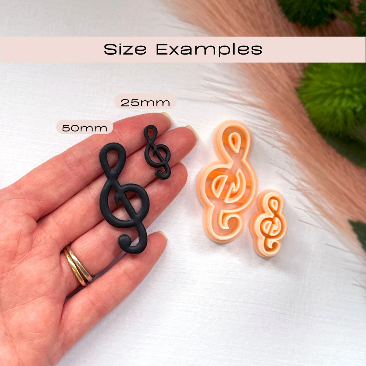 Two treble clef-shaped clay cutouts shown on a hand for scale, with labels reading 25mm and 50mm, plus two larger peach treble clef and a smaller orange treble clef cutout on a white surface. Text at the top says 'Size Examples'.