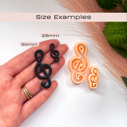 Two treble clef-shaped clay cutouts shown on a hand for scale, with labels reading 25mm and 50mm, plus two larger peach treble clef and a smaller orange treble clef cutout on a white surface. Text at the top says 'Size Examples'.