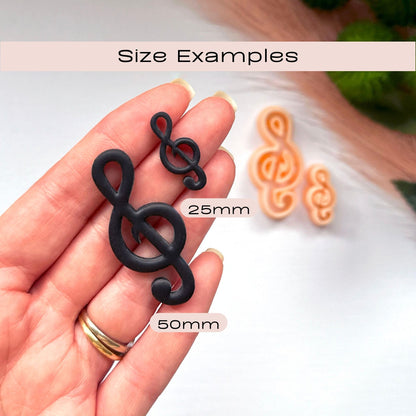 A hand holds two black treble clef-shaped items labeled 25mm and 50mm, with two peach-colored treble clefs blurred in the background. Text at the top reads 'Size Examples'.