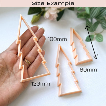 Three peach-colored plastic cookie clay cutters shaped like stylised Christmas trees, with one being held in a hand to show scale on a white surface with soft floral background. Great for ornament making. Size example: 120 mm, 100 mm, 80 mm written.