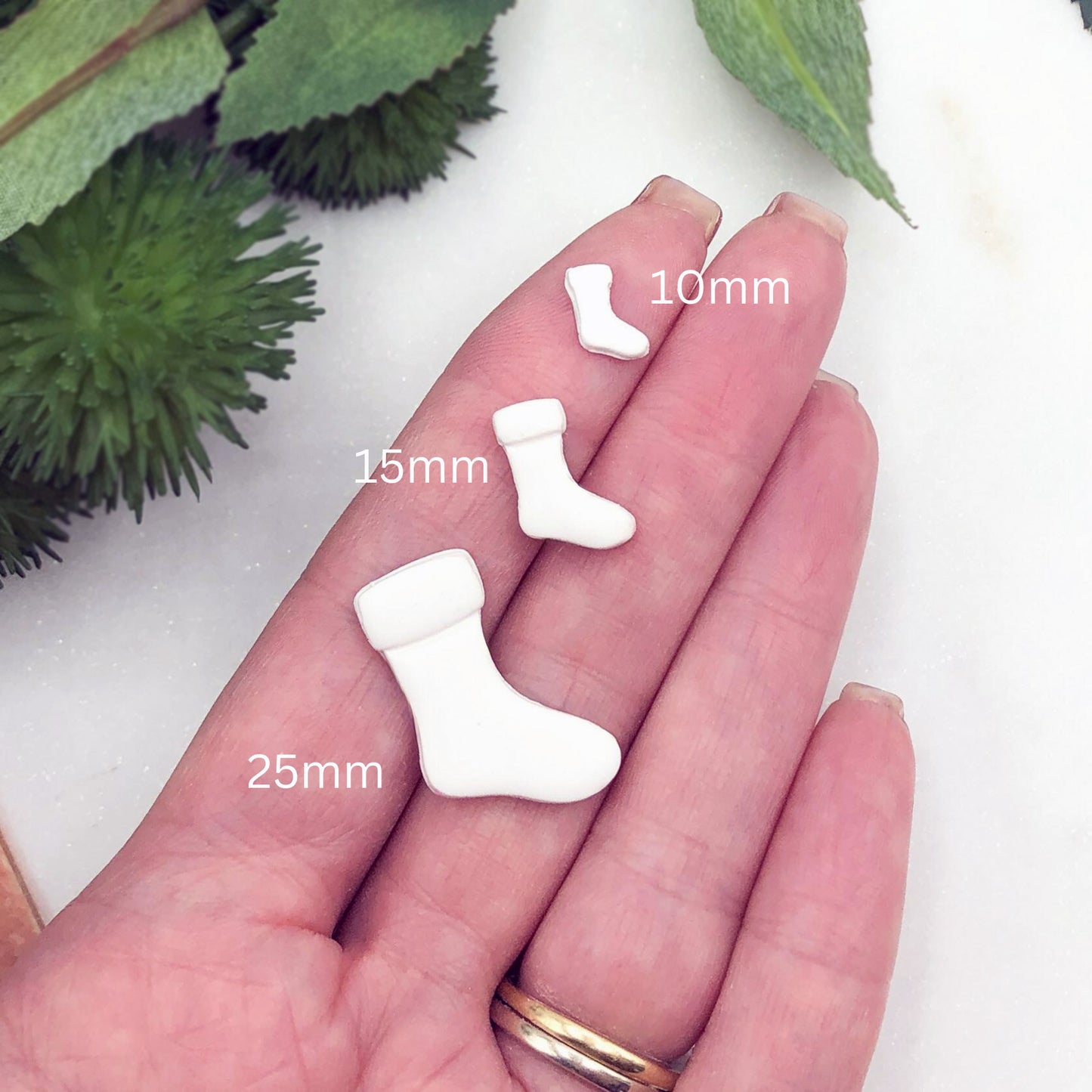 Three small white sock-shaped charms  displayed on a person's palm, labelled with sizes 10mm, 15mm, and 25mm. Green leaves are visible in the background.