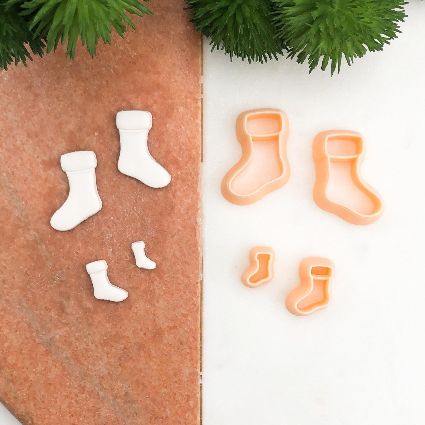 A set of miniature sock-shaped cookie cutters in peach and matching white fondant cutouts, arranged on a split terracotta and white background with green pine branches at the top.